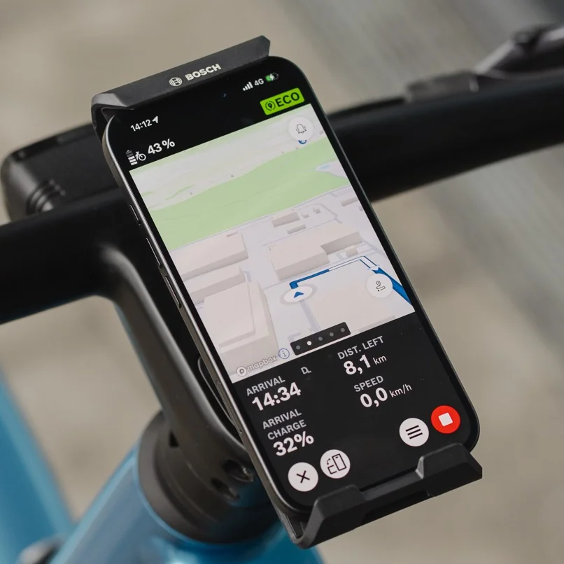Close-up of a smartphone attached to the handlebars in the Bosch eBike SmartphoneGrip Vertical, used as an eBike display for navigation. The eBike Flow app shows a colorful road map with a blue route line, an upcoming left turn, and the remaining distance and arrival time at the bottom of the screen.