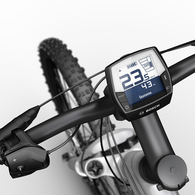 Un écran Intuvia est fixé au guidon d’un e-MTB noir et argent. Il affiche ta vitesse, le mode de conduite, le niveau de batterie et le rapport sélectionné.