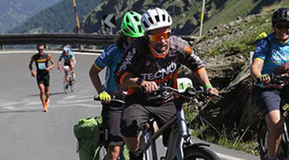 Un uomo e una donna pedalano su una bici tandem lungo una strada tra le rocce, con altri ciclisti di eBike visibili sullo sfondo.
