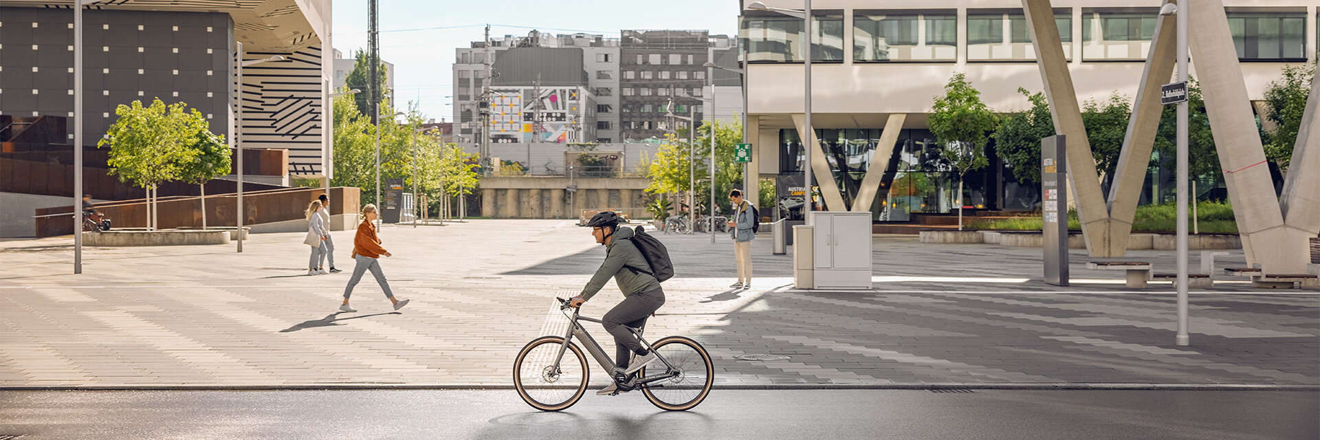 An eBike rider drives down a pathlong, in the background you can see buildings and pedestrians.
