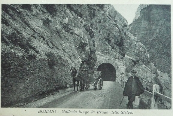 Scatto in bianco e nero di una carrozza trainata da cavalli e due uomini in piedi davanti a una montagna con un tunnel. La didascalia recita: "BORMIO - Galleria lungo la strada dello Stelvio".