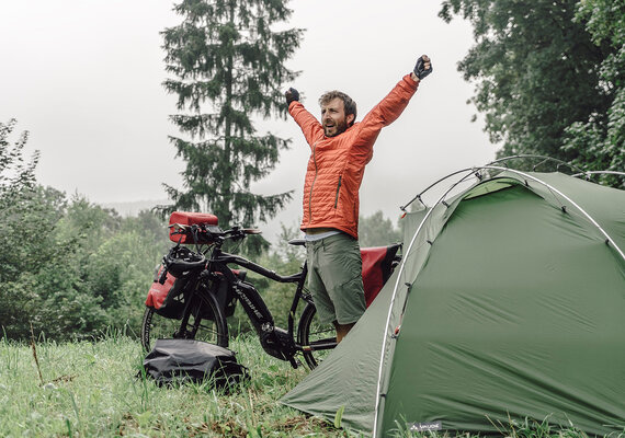 Der Fotograf Maximilian Semsch steht zwischen seinem eBike und Zelt und streckt seine Arme in die Höhe