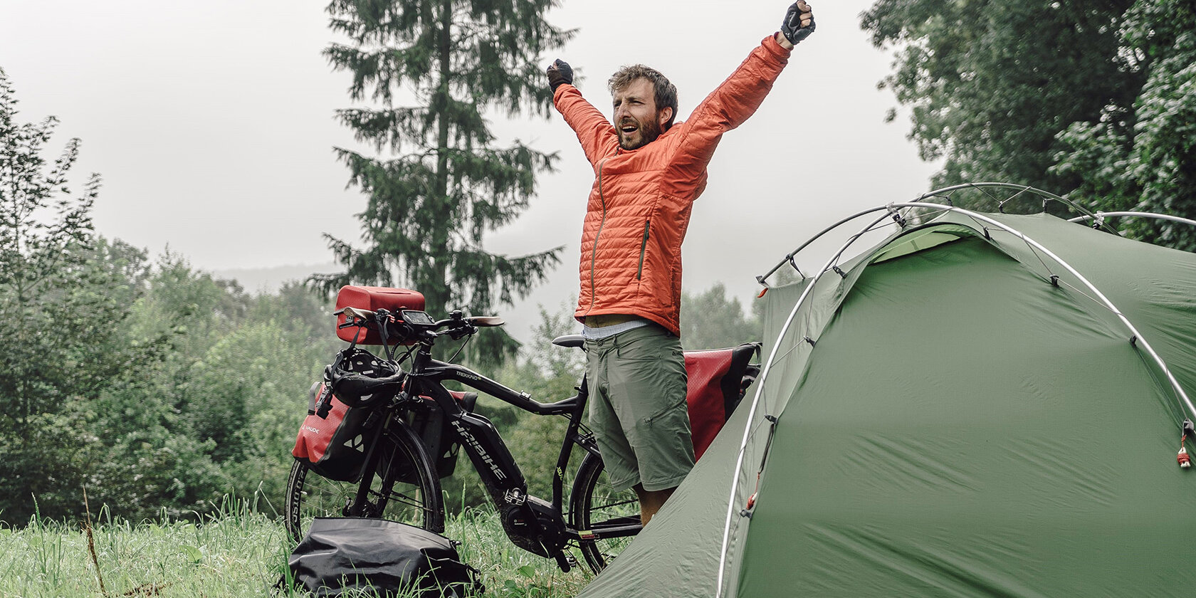 Der Fotograf Maximilian Semsch steht zwischen seinem eBike und Zelt und streckt seine Arme in die Höhe