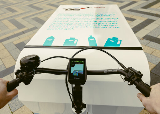 Beeld vanuit het perspectief van de bestuurder van een bakfiets met een laadbak als laadruimte. Je ziet het stuur met display, en een witte laadbox waarop in blauwe letters iets is gedrukt.