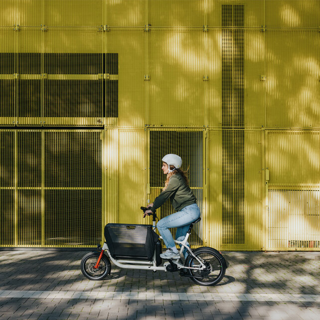Une femme avec un casque roule sur un VAE Cargo le long d’un bâtiment jaune.