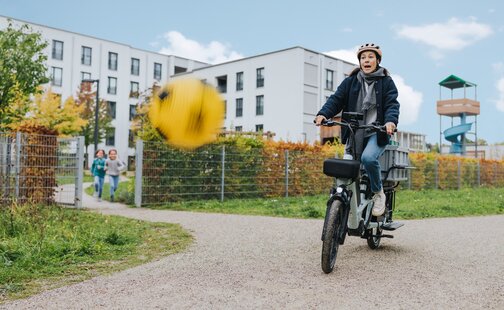 A woman suddenly brakes on her eCargo bike when a yellow ball bounces into the air in front of her. She looks startled.