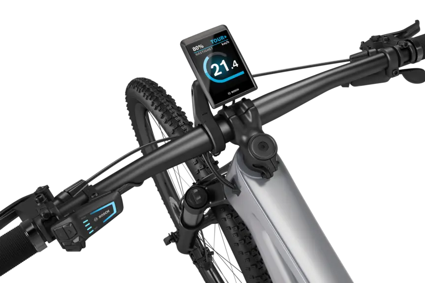 En sølvfarget eMountainbike med svart styre plassert mot en hvit bakgrunn. På styret er en Kiox 500 montert. Displayet viser hastigheten i km/t, batterinivået og kjøremodusen.