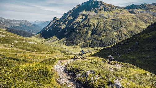 Deux cyclistes électriques parcourent la route Heckmair à travers les Alpes, entourés de paysages montagneux et de sentiers escarpés.