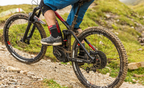 Nahaufnahme eines mit Matsch bedeckten eBikes, welches von einer Person über einen erdigen und steinigen  Weg zwischen Gras gefahren wird.