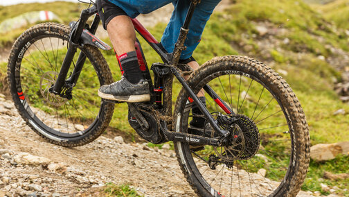 Close-up of an [eBike] covered in [M]ud, which is being ridden by a person along an earthy and stony path between grass.