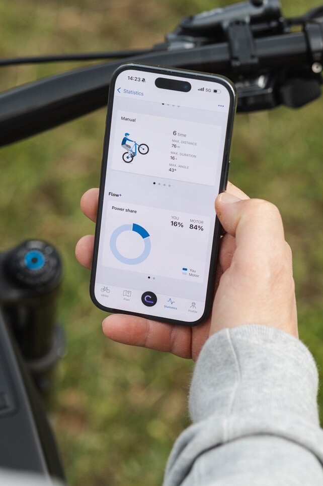Gros plan sur une main tenant un smartphone au-dessus d’un guidon VAE. Sur l’écran, les statistiques de figures pour les manuelles ainsi que la part de performance du pilote et du moteur sont affichées dans l’application eBike Flow.