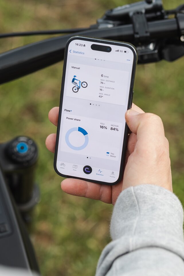Close-up of a hand holding a smartphone above an eBike handlebar. The display shows trick statistics for manuals and the performance share between the rider and motor in the eBike Flow app.