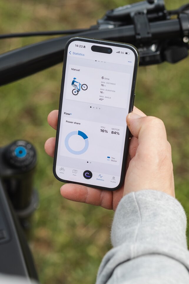 Close-up of a hand holding a smartphone above an eBike handlebar. The display shows trick statistics for manuals and the performance share between the rider and motor in the eBike Flow app.