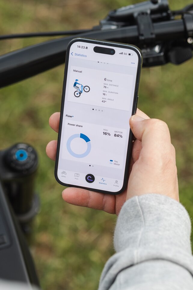 Close-up of a hand holding a smartphone above an eBike handlebar. The display shows trick statistics for manuals and the performance share between the rider and motor in the eBike Flow app.