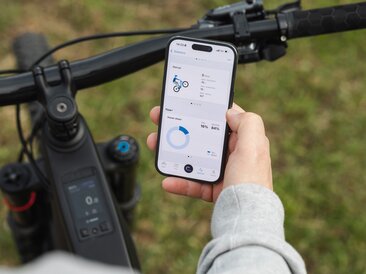 Gros plan sur une main tenant un smartphone au-dessus d’un guidon VAE. Sur l’écran, les statistiques de figures pour les manuelles ainsi que la part de performance du pilote et du moteur sont affichées dans l’application eBike Flow.