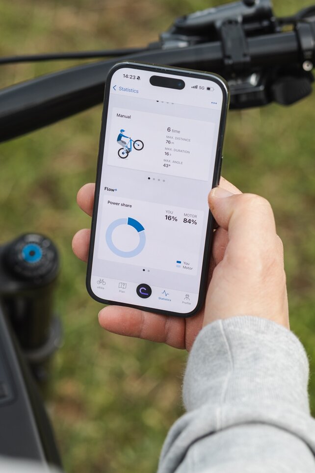 Primo piano di una mano che tiene uno smartphone sopra un manubrio eBike. Sul display, vengono visualizzate nel app eBike Flow le statistiche delle aperture per i manuali e la quota di prestazione del pilota e del motore.