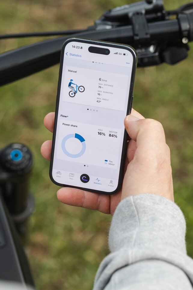 Gros plan sur une main tenant un smartphone au-dessus d’un guidon VAE. Sur l’écran, les statistiques de figures pour les manuelles ainsi que la part de performance du pilote et du moteur sont affichées dans l’application eBike Flow.