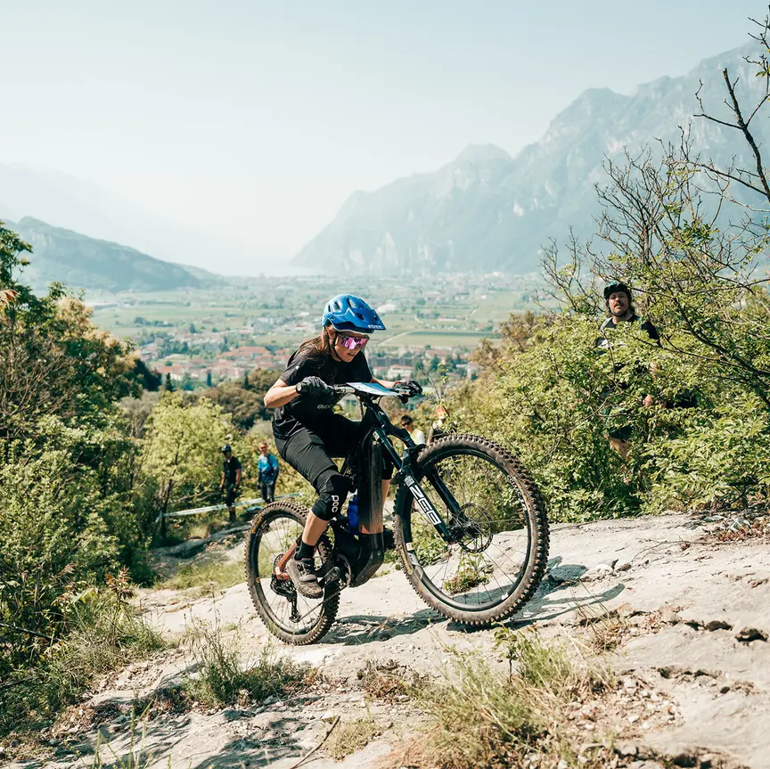Une cycliste gravit des rochers sur son VTT AE. À l’arrière-plan, une vallée entourée de montagnes. Plusieurs villages se trouvent dans la vallée.