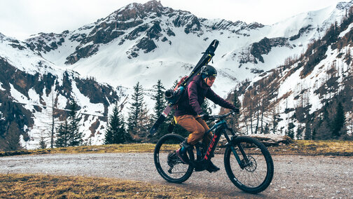 Un conducteur de VAE roule sur un chemin de gravier avec un équipement de ski accroché à son sac à dos. À l’arrière-plan, on aperçoit des montagnes enneigées.