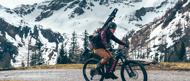 Un conducteur de VAE roule sur un chemin de gravier avec un équipement de ski accroché à son sac à dos. À l’arrière-plan, on aperçoit des montagnes enneigées.