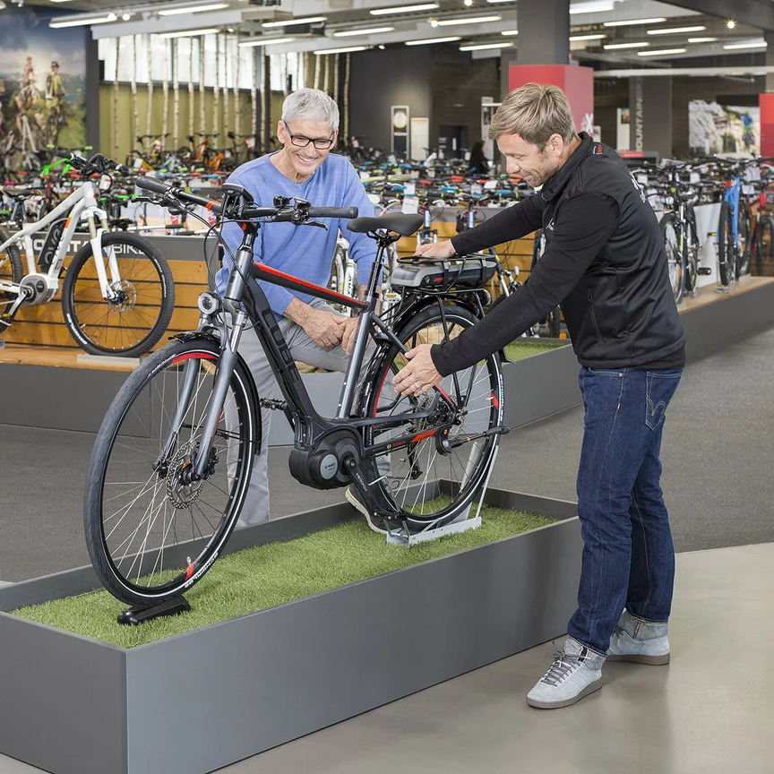 Konzultant Bosch eBike / elektrokolo stojí s mužem v obchodě s jízdními koly před výstavním kolem a vše mu podrobně vysvětluje.