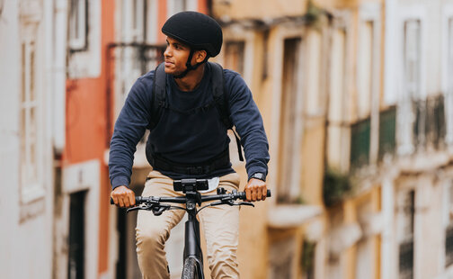 A man is riding his eBike through an alley, glancing to the side. A smartphone is attached to the eBike, with houses visible in the background.