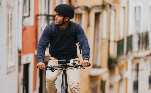 Mann fährt mit seinem eBike durch eine Gasse und schaut leicht zur Seite. Ein Smartphone ist am eBike befestigt. Im Hintergrund sind Häuser zu sehen.