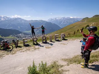 Four individuals are depicted in the image. A man is using his smartphone to take a photo of two women standing on large wooden letters forming the word "Verbier". In the background, mountains are visible.