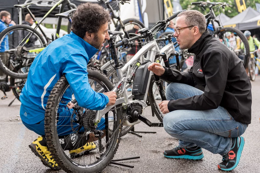 An eBike advisor and a man kneel in front of an eBike, talking together. Outside, more eBikes are visible in the background.