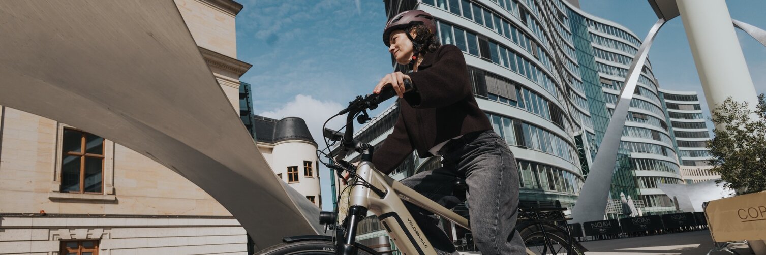 Une personne à vélo en VAE traverse un paysage urbain saisissant. La prise de vue dynamique en contre-plongée met en valeur le contraste entre un bâtiment en verre courbé ultramoderne et une architecture historique sous un ciel bleu.