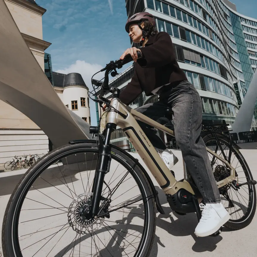 Eine Radfahrerin auf einem eBike fährt durch eine beeindruckende Stadtkulisse. Die dynamische Aufnahme aus der Froschperspektive fängt den Kontrast zwischen einem hochmodernen, geschwungenen Glasgebäude und historischer Architektur unter blauem Himmel ein. 