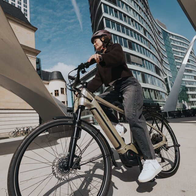 Cyklista na eBike projíždí působivou městskou scenérií. Dynamický záběr z nadhledu žabí perspektivy vystihuje kontrast mezi moderní zakřivenou skleněnou budovou a historickou architekturou pod modrou oblohou.