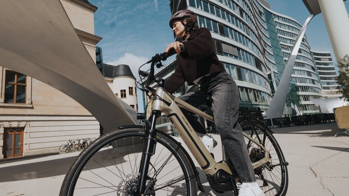 A cyclist on an eBike rides through an impressive cityscape. The dynamic, low-angle shot highlights the contrast between a state-of-the-art curved glass building and historic architecture under blue skies.