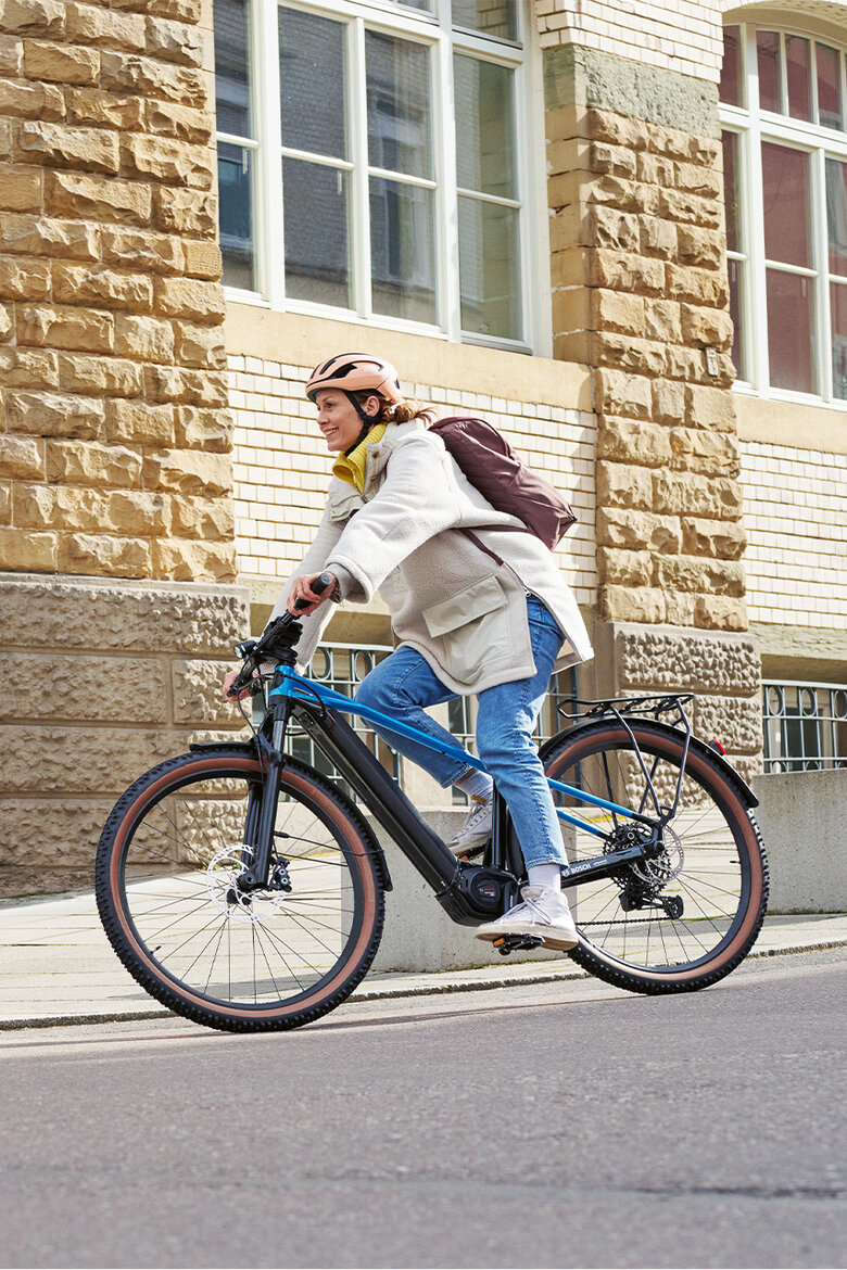 Eine Frau fährt lächelnd mit ihrem eBike durch die Stadt. Im Hintergrund ist ein Gebäude zu sehen.