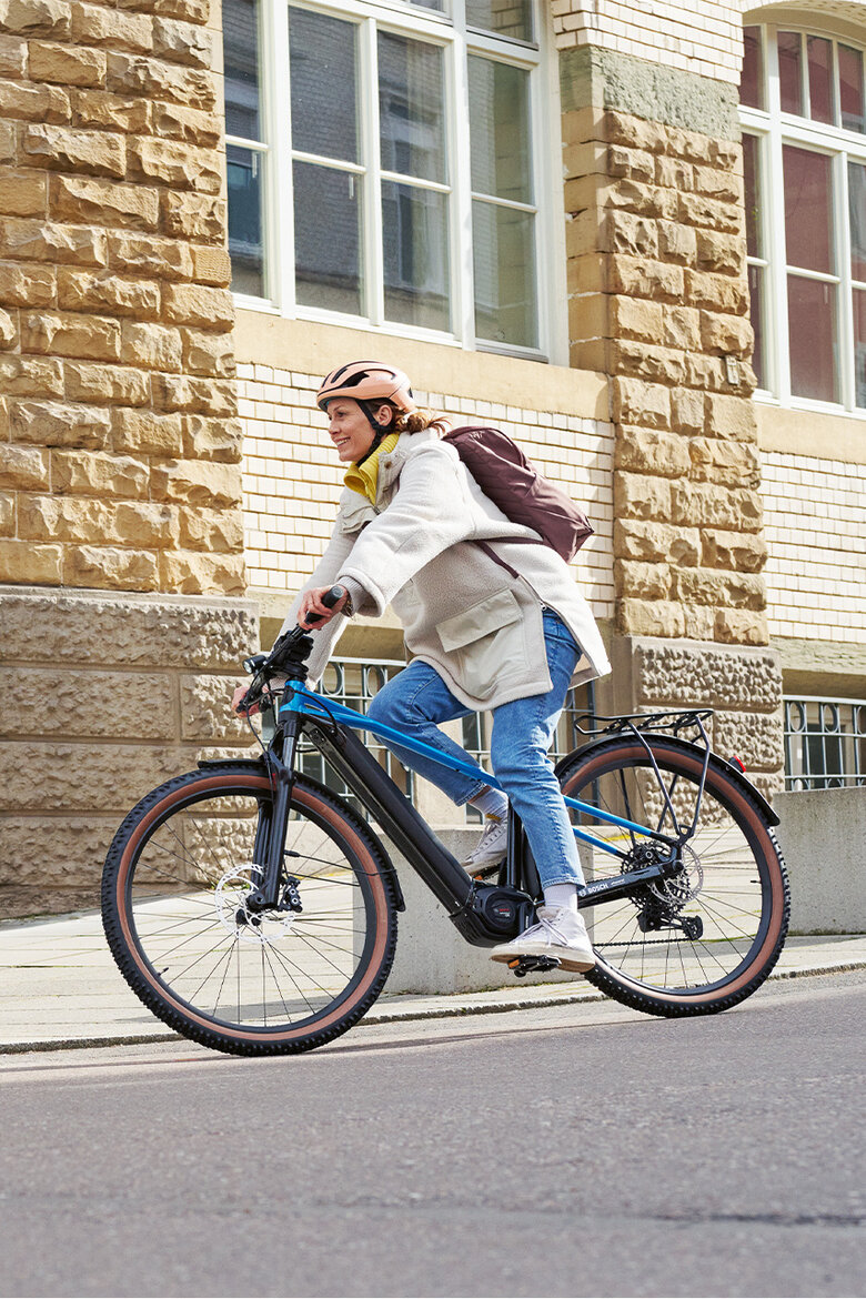 A smiling woman rides her [eBike] through the [city]. A building can be seen in the background.
