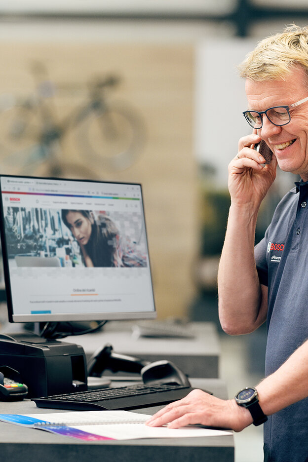Esperto di [eBike] è in piedi alla sua scrivania nel negozio di biciclette. Sul computer è aperto il sito web di Bosch mentre è al telefono con il [Kundenservice] di Bosch.