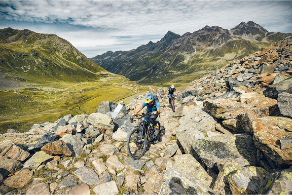 Dos ciclistas de eMountainbike atraviesan las rocas en las montañas, con colinas y montañas de fondo.