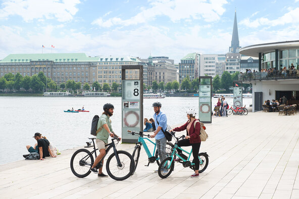 Drei eBiker mit ihren Pedelcs, die sich in der Innenstadt Hamburgs an einem Flussufer unterhalten