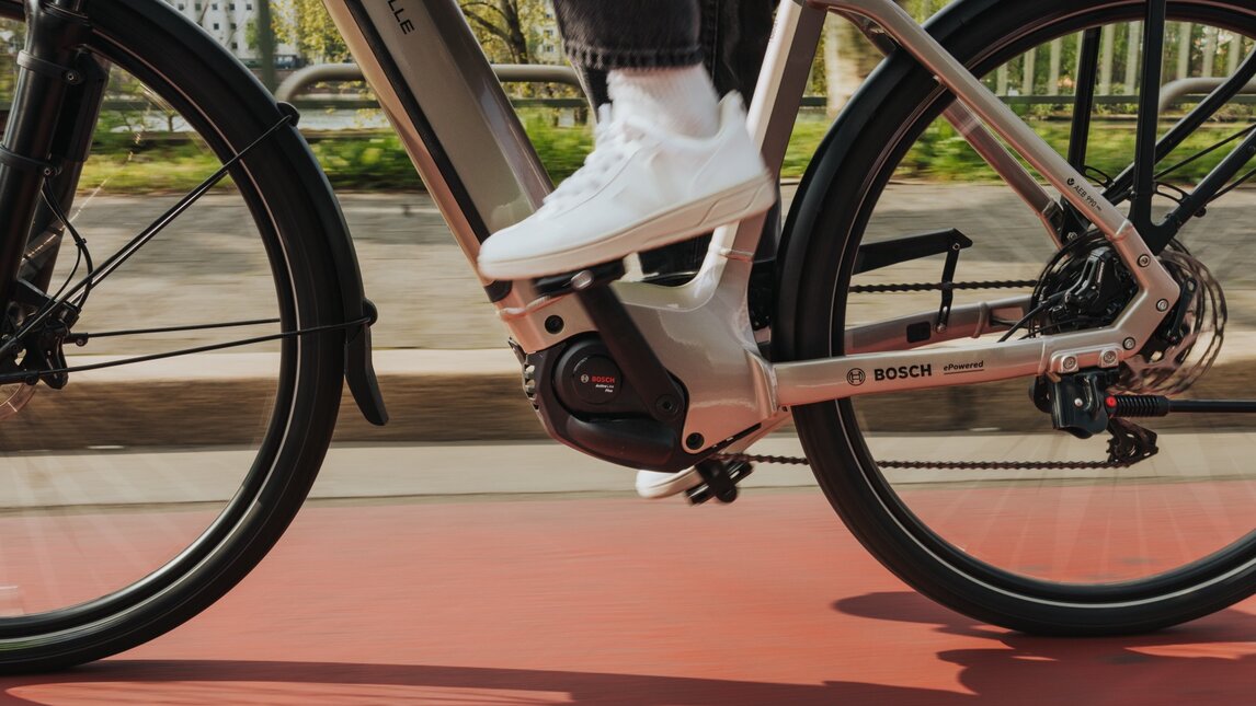Shot of an eBike riding on a red bike lane. The focus is on the central Bosch motor Active Line Plus and the foot in a white sneaker on the pedal. The motion blur of the ground and the distinct shadows cast create a strong sense of speed on a sunny day.