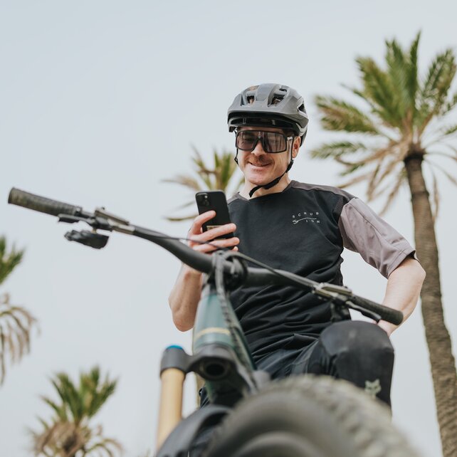 Ein lächelnder eMountainbiker mit Helm und Schutzbrille macht eine Pause auf seinem eBike und schaut auf sein Smartphone. Im Hintergrund sind Palmen vor einem hellen Himmel zu sehen. Die Aufnahme ist aus der Froschperspektive, über das Vorderrad des Fahrrads hinweg, fotografiert.