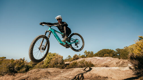 An eBike rider jumps his turquoise mountain bike over an earthy hill against a blue sky.