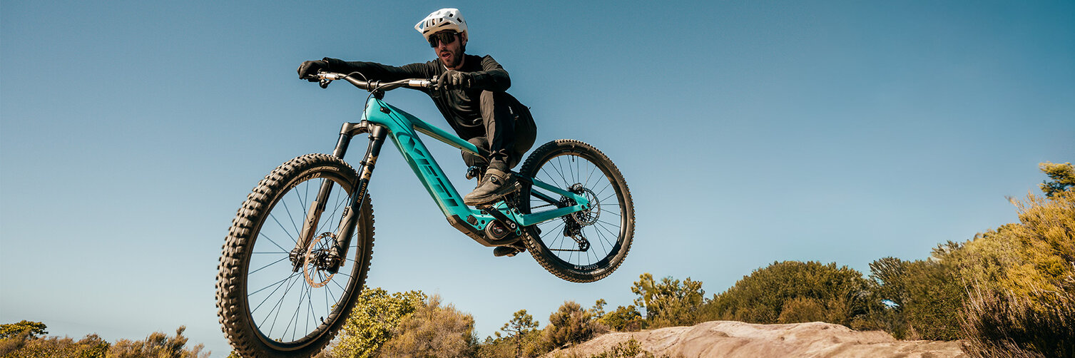 An eBike rider jumps his turquoise mountain bike over an earthy hill against a blue sky.