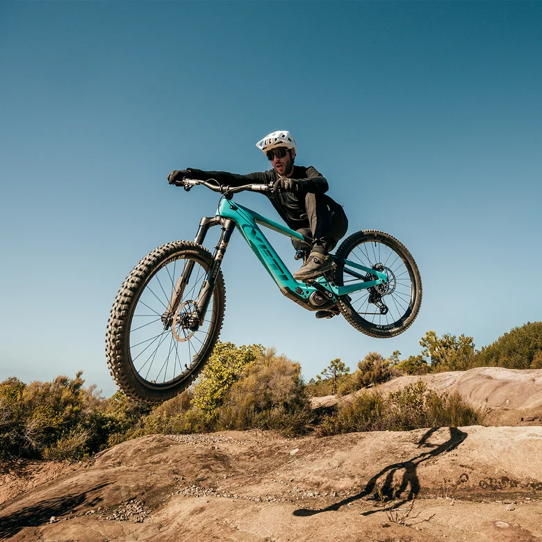 An eBike rider jumps his turquoise mountain bike over an earthy hill against a blue sky.