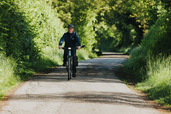 Frontalansicht eines lächelnden Mannes, der mit seinem eBike einen Fahrradweg im Grünen entlangfährt.