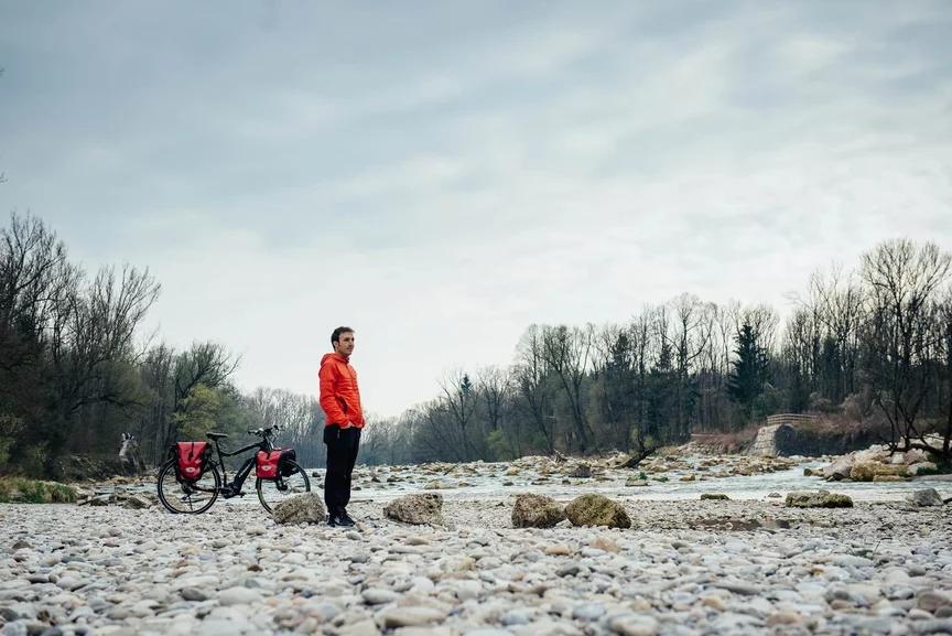 Muž v červené bundě stojí v kamenitém korytě řeky, za ním je jeho eBike. Na okraji břehu rostou stromy a mezi kameny je vidět voda.