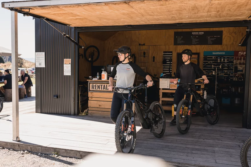 Two eMountain bike riders push their eBikes out of a shed, where a reception area marked 'Rental' is visible.