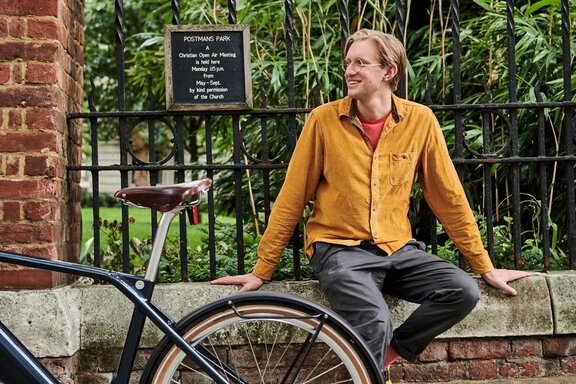 Architekt Edward Crooks siedzi na ceglanym murze, przed nim stoi jego czarne eBike. W tle rozciąga się park z zielonym bambusem i łąką.