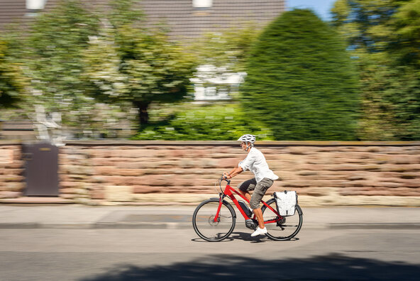 Frau mit eBike-Helm fährt auf rotem Pedelec an einem Haus mit großem Garten vorbei