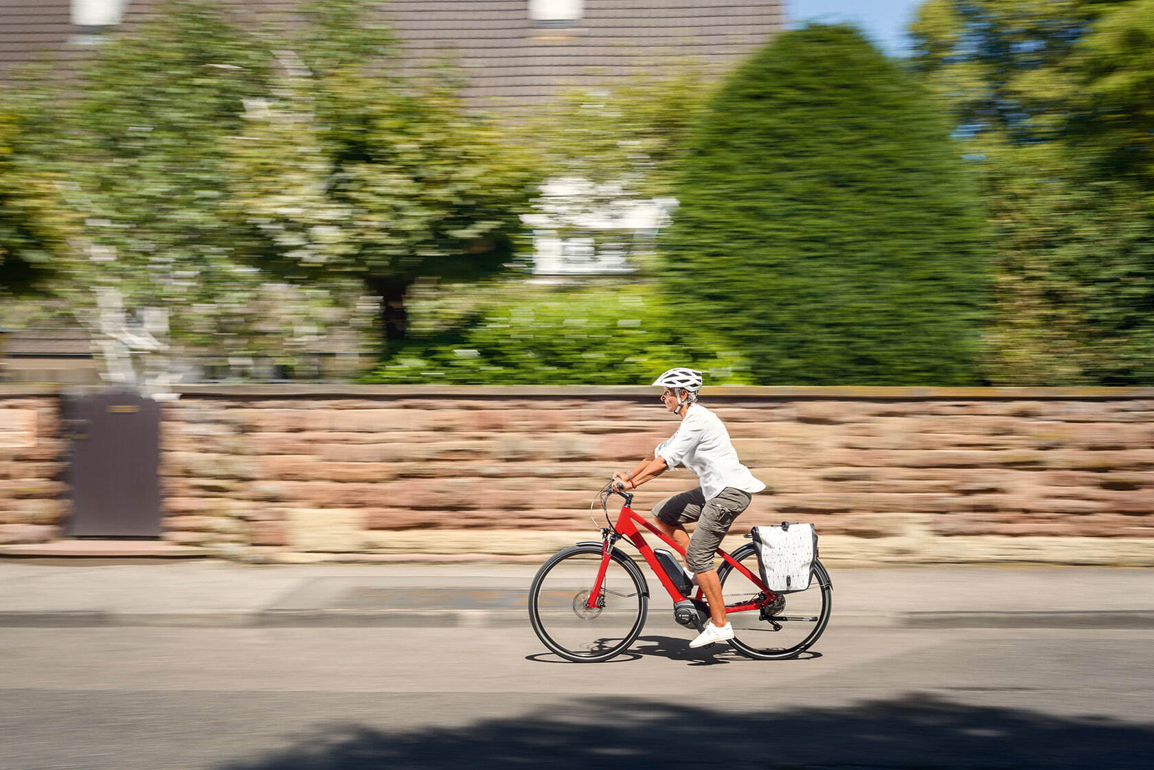 Une femme (eBike) utilisatrice conduit sur un (eBike) rouge le long d'une route, à côté de laquelle se trouve un mur de pierre avec une porte séparant un jardin avec des arbres et des buissons de la route.