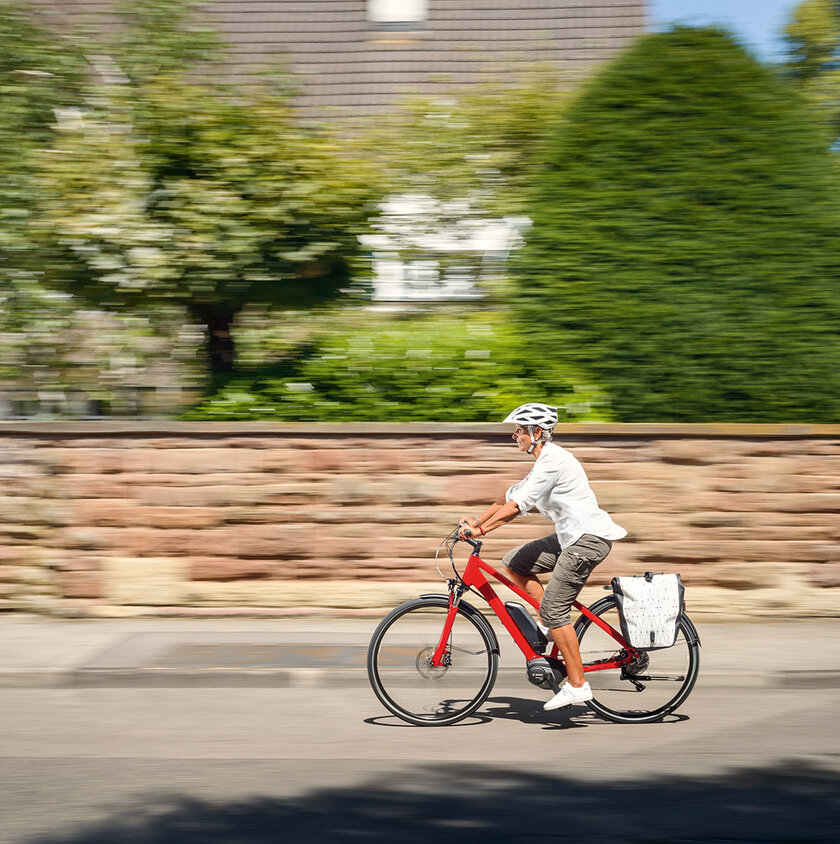 Frau mit eBike-Helm fährt auf rotem Pedelec an einem Haus mit großem Garten vorbei
